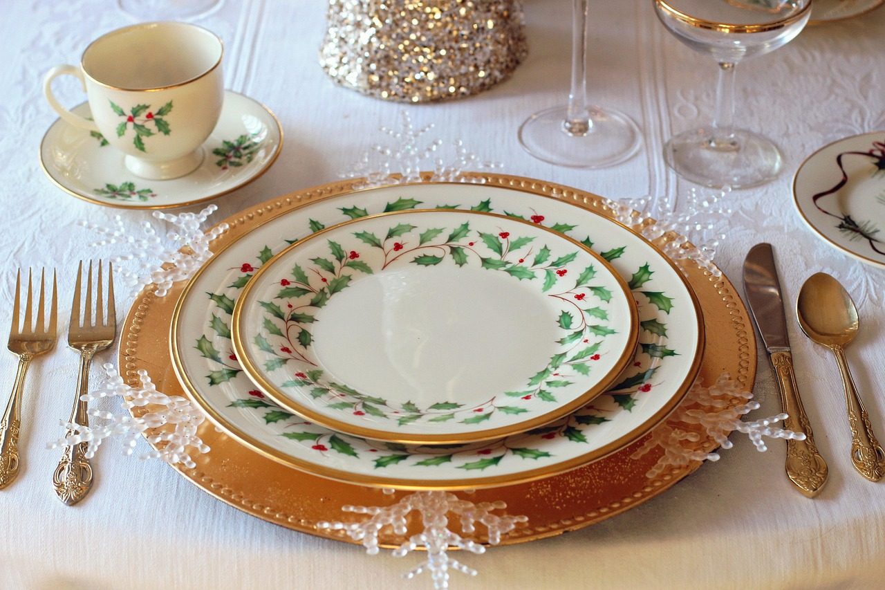 Table de fête dressée pour Noël avec une vaisselle blanche décorée de houx, des couverts dorés et un service élégant sur nappe blanche